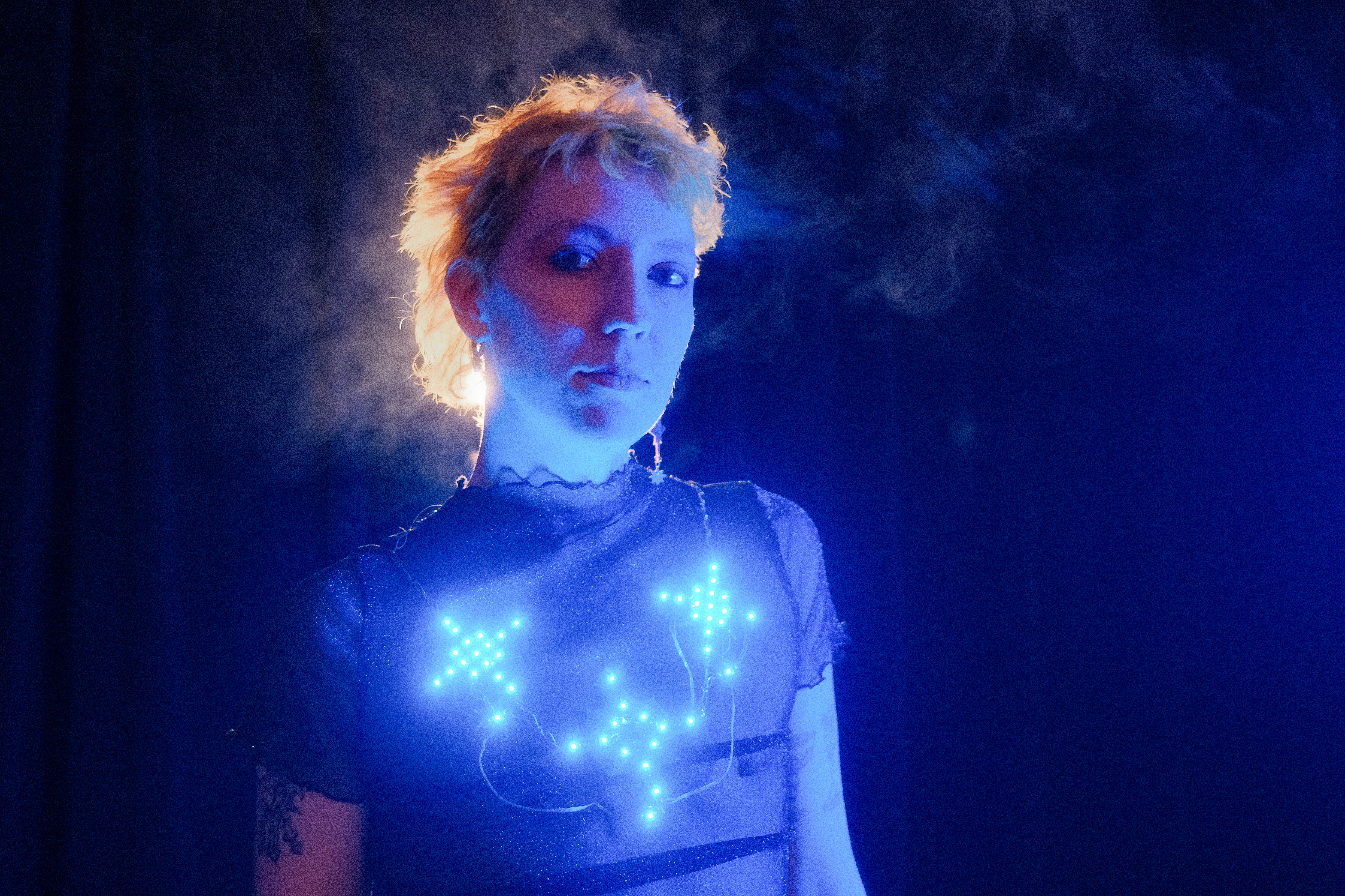 Photo of Kay against a dark background, wearing a bright blue light up neacklace in the shape of stars.